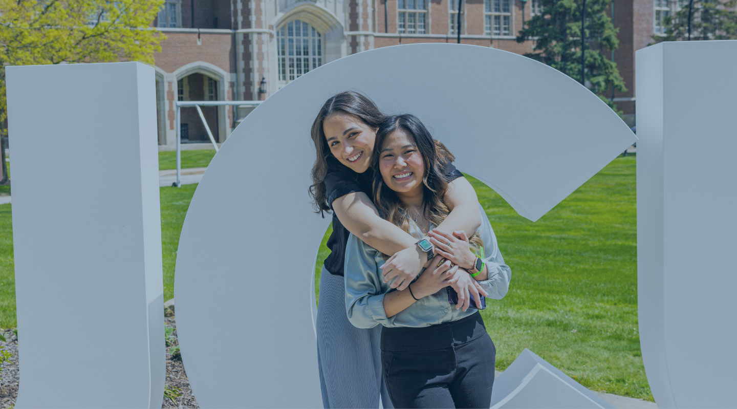 Two girls hugging in front of white cement letters spelling JCU