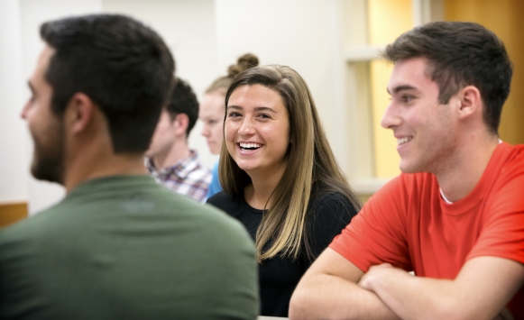 Three students sit in a classroom. One female student smiles at the camera. 