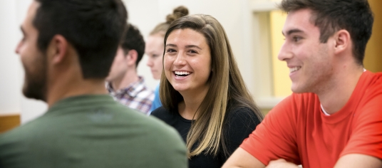 Three students sit in a classroom. One female student smiles at the camera. 